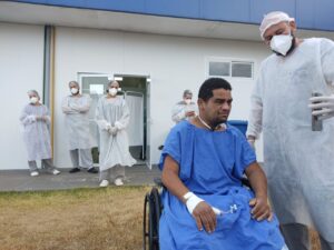 Homem está sentado numa cadeira de rodas vestido de azul enquanto um outro homem está de pé a sua direita, vestido de branco, máscara e touca hospitalar branca.
