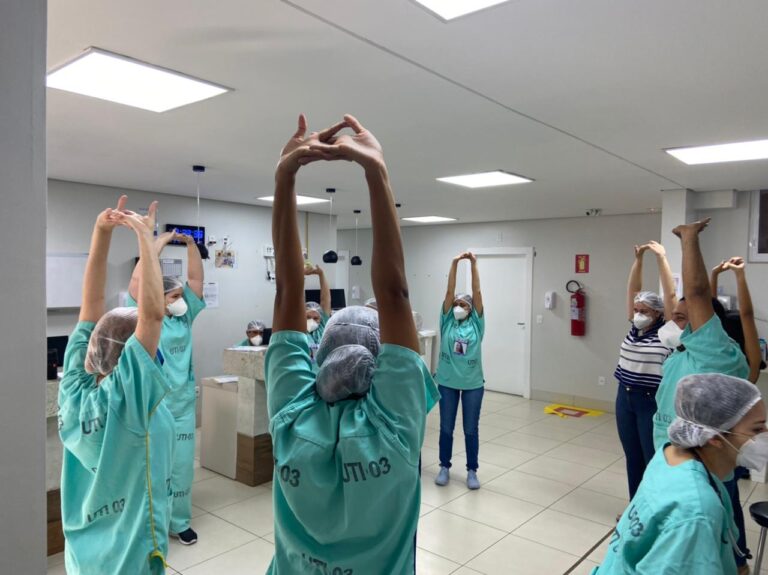 Pessoas com fardamento na cor verde. Elas estão umas ao lado das outras, formando um círculo. Todas estão com os braços esticados para cima, fazendo alongamento.