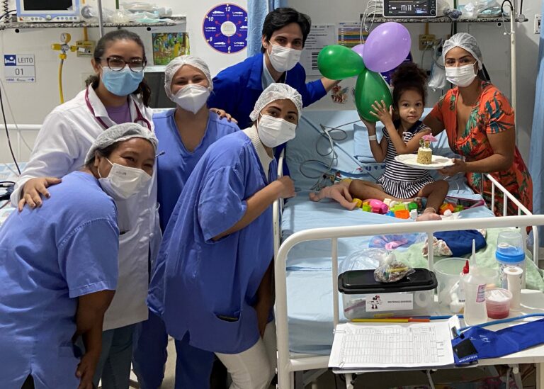 Cinco pessoas com jalecos brancos e azuis estão ao lado esquerdo de um leito. Sentada no leito está uma criança com bolo na mão. Do lado direto do leito hospitalar está uma mulher de máscara e vestido colorido. Ao fundo, aparece balões de aniversário.