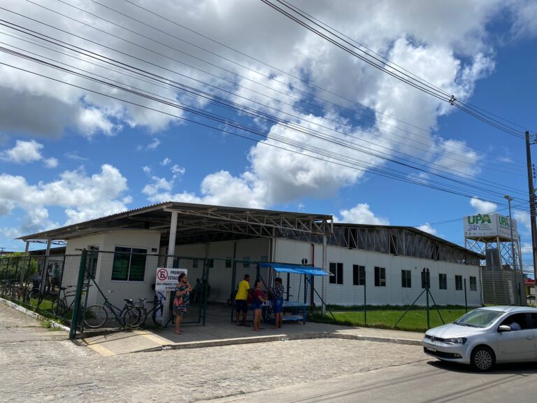 Foto de um dos acessos à UPA Benedito Bentes. Na imagem aparece parte do prédio branco, de único pavimento, várias janelas e pessoas próximas à grande. Há um carro estacionado em frente ao prédio. A rua está vazia.