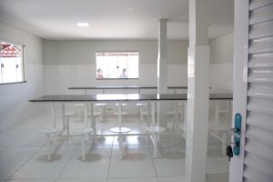 Imagem geral do refeitório. Paredes e piso branco com uma mesa no centro do salão..